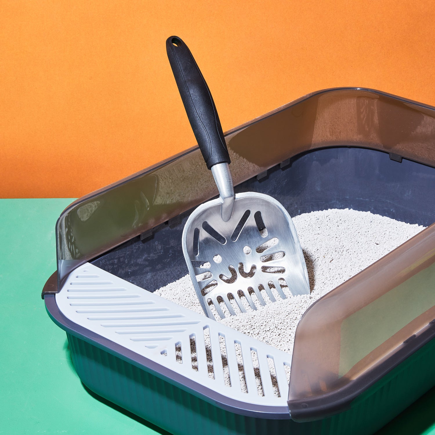 Our award-winning clay cat litter in our grey plastic tray with a cat-faced scoop and an orange and green background.
